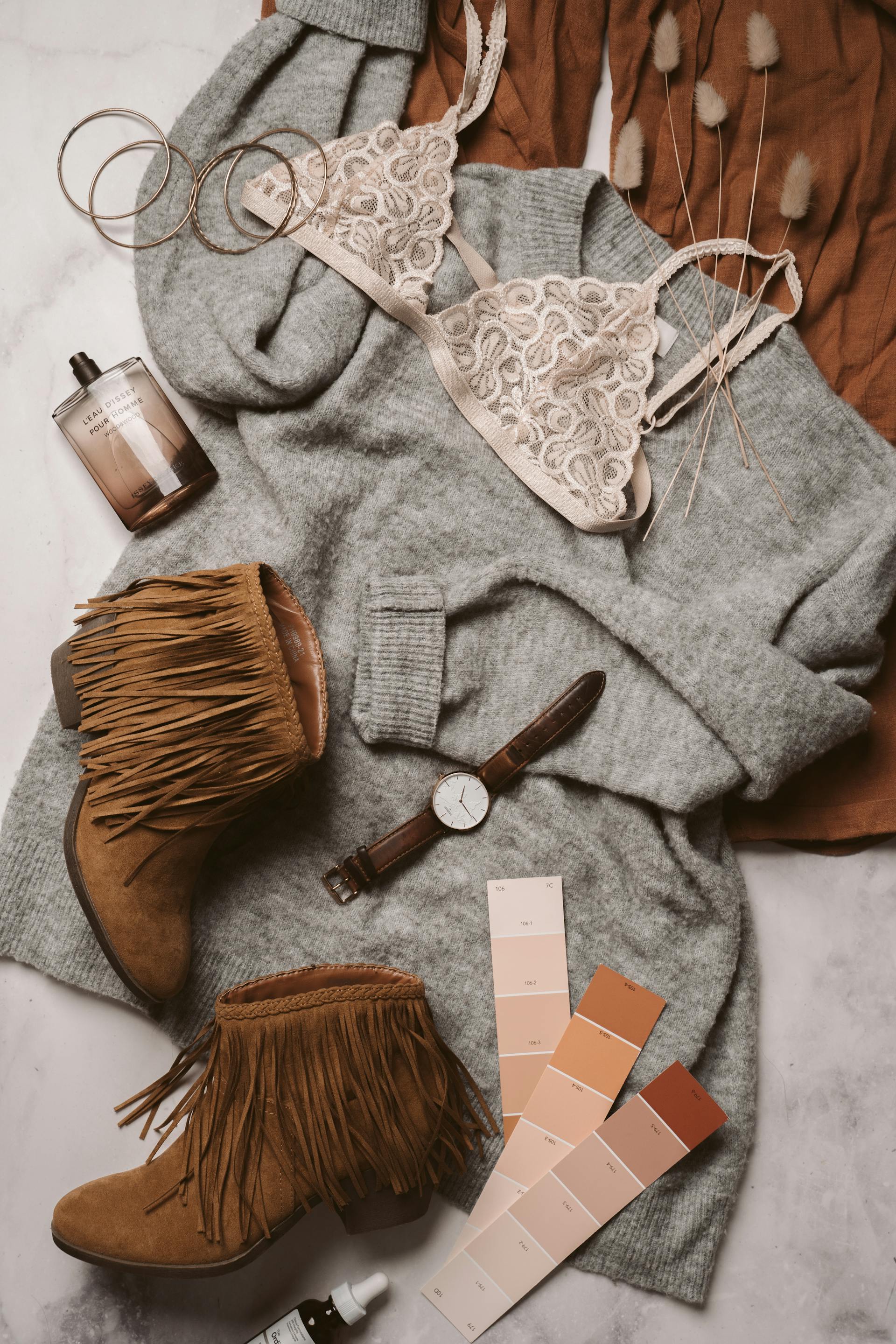 Editorial flat lay — grey sweater, fringe boots, lace bralette, perfume, leather watch, color swatches in warm neutrals.