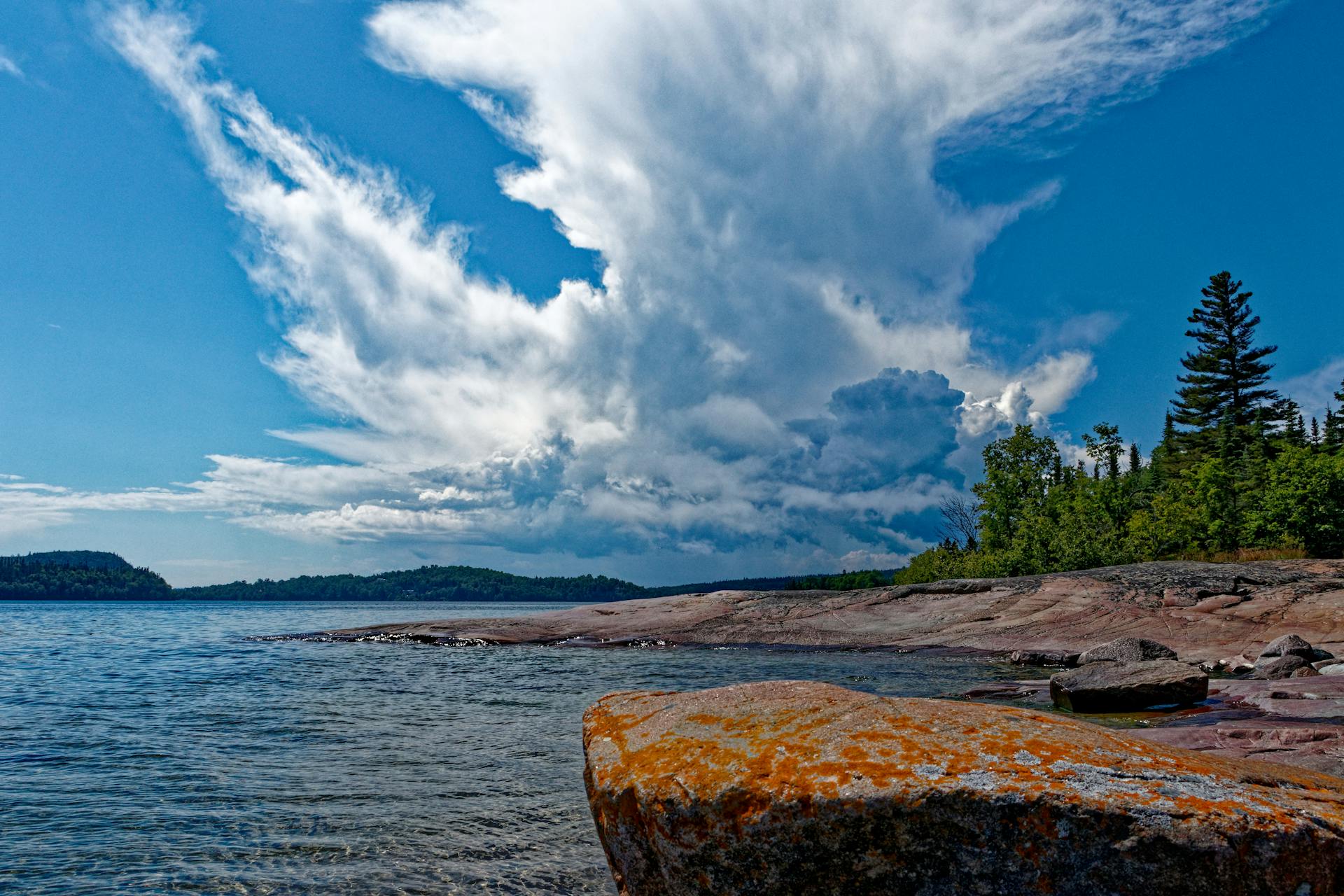 Lake Superior shoreline near Marquette, MI — iron-orange lichen on a sun-warmed boulder, pines on the bank, dramatic UP sky overhead.